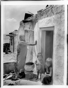 Hopi Indian mother whitewashing her adobe dwelling in the doorway of which are two children, ca.1900