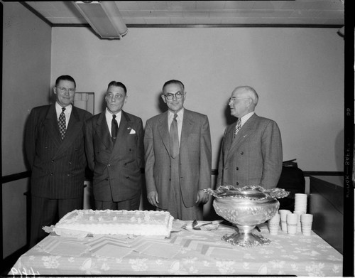 Four men standing in front of a large sheet cake and punch bowl
