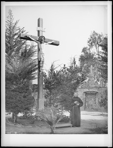 Priest standing next to a tall wooden crucifix in the cemetery at Mission Santa Barbara, 1898