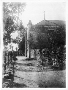 View of the east end of Mission San Gabriel Arcangel from the walk to the cemetery, ca.1900