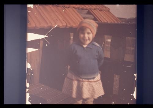 Photograph of child in hat standing on porch and smiling