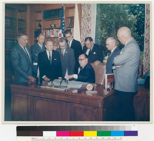 Governor Edmund G. Brown, seated at his desk, pointing to a map, surrounded by men