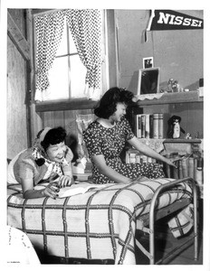 "Oko Murata, left, and Esther Naito, former office workers, evacuees now living at the Manzanar relocation center, proudly smile at the personal library they have established in their camp dormitory quarters."--caption on photograph