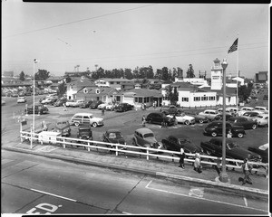 Exterior view of the Farmers Market, showing Gilmore Field and parked automobiles, ca.1953