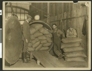 Three workers inside a factory, surrounded by large sacks, ca.1930