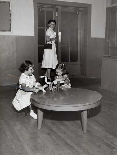 Two little girls sitting at a table playing while a woman watches