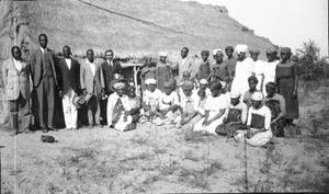 Swiss missionary and African people in front of a building with a thatched roof, Cheu, Mozambique
