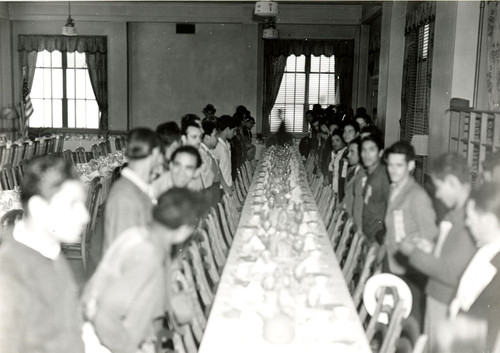 Dining hall with long tables and newly arrived Mexican workers standing at the chairs