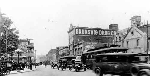 View of Main Street, includes church, and Brunswig Building (Brunswig Drug Company) on the right and the Plaza and Pico House on the left. Heavy traffic, antique cars and trolley cars