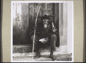 A beggar who lives near Kutschuk. He holds the beggar's staff and rice-bowl - or beggar's bowl - in his hands and waits patiently till the schoolchildren, or perhaps the missionary, has eaten, so that he can be given whatever rice is left over