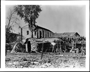 View of the Mission San Gabriel from the east end, showing the first bell tower and the last of the Mission Indians settlement, ca.1900