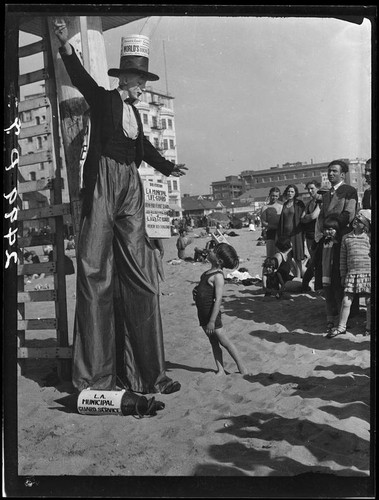 Clown on stilts, advertising dance marathon, with child on beach, Santa Monica, 1928