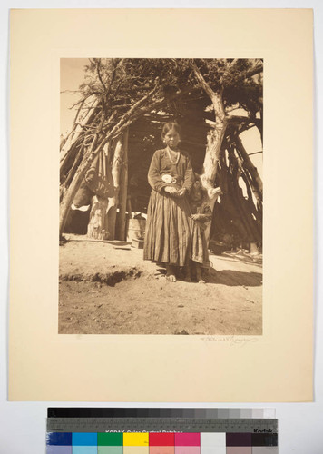 Navajo girl and a younger child standing in front of a wood and brush shelter