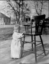Young toddler standing at a high chair outside in a yard, 1930s