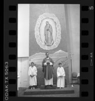 Father Luis Olivares standing below Virgin of Guadalupe icon, Los Angeles, Calif., 1986
