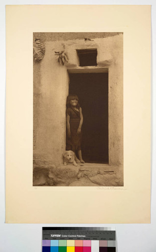Hopi girl standing in a doorway of an adobe building, with a dog at her feet