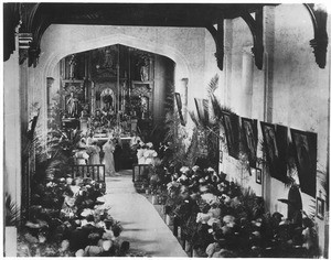 Wedding consisting of wedding principals, attendants and guests standing before the ornate alter in the San Gabriel Mission, ca.1900