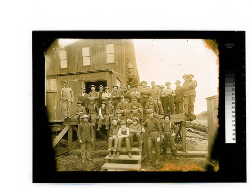 [Large group of men & boys pose by an industrial building]