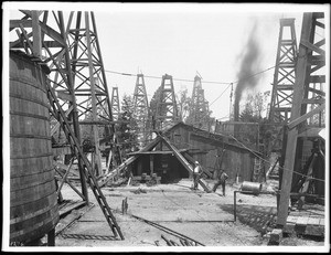 Two men standing near a wooden shed in the midst of dozens of oil derricks in a Los Angeles oil field