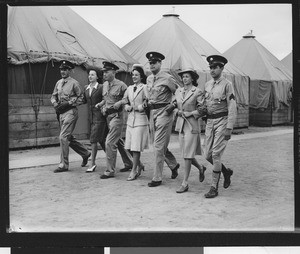 Row of seven people in uniform walking arm in arm past a row of tents during World War II, ca.1939