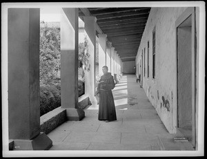 Monk with a book standing in the inner corridor and patio of Mission Santa Barbara, 1898