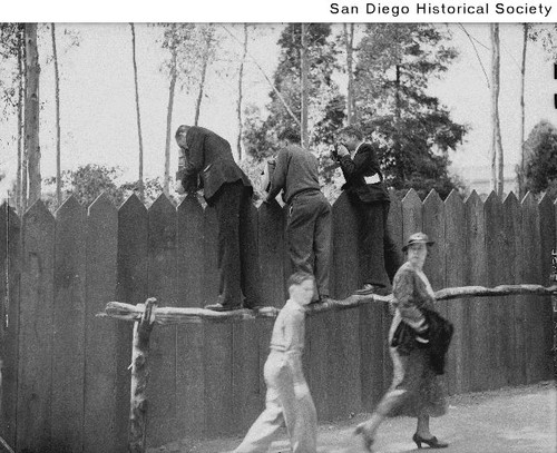 Three men with cameras standing on a handrail looking over a fence as a woman and young man walk by