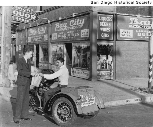 A man handing a sack to a Bay City Liquor delivery driver who is seated on a motorized delivery tricycle outside Bay City Liquor