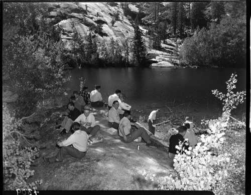15 men eating lunch and drinking beer on a rock beside a lake in the Big Creek area