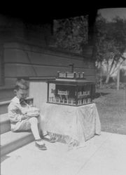 Child's birthday party with a young boy seated on a front step next to a table with a fort-like toy, about 1920-30