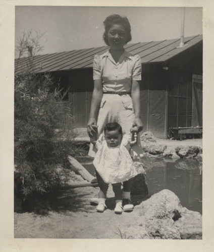 Woman and infant stand in front of pond and barrack at Poston incarceration camp