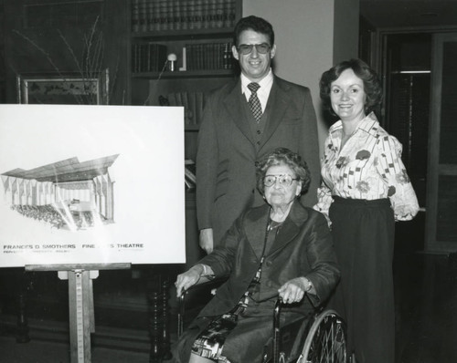 Mrs. Smothers seated in a wheel chair next to a sketch of the proposed Smothers Theatre; standing behind her are Dr. Larry Hornbaker and Carol