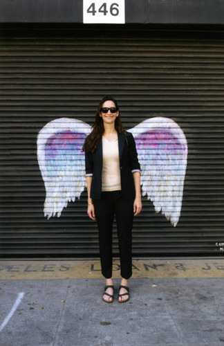 Unidentified woman in a black blazer and sunglasses posing in front of a mural depicting angel wings