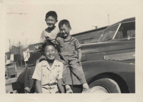 Three smiling boys sit on a car at Poston incarceration camp