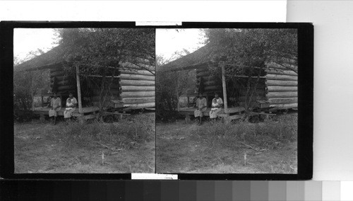 U.S. Indian Reservation. You are now looking at Mrs. Florence Bullock, her daughter & grandchild sitting on the porch of the old log hut that was their home before the government took charge of these people. They are now using this for a smoke-house. I went down to the home of Charles Anderson who is the chief of this reservation for the purpose of photographing him & found him sick in bed & not expected to live. He has been sick about one and a half yrs (1 1/2) & this was the closest I came to finding an Indian family at home as everyone else was engaged in their work in the fields