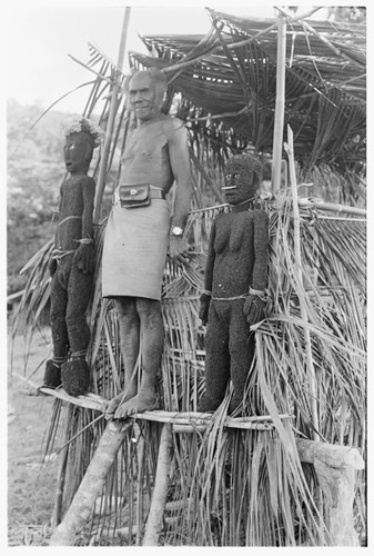 Folofo'u, wearing old police belt, on speaking platform with fernwood 'ea figures made by Arimae of Furi'ilae for the opening of the Kwaio Cultural Centre in August 1979