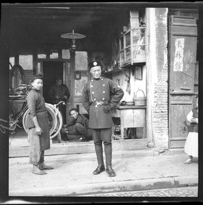 Man in a military police uniform standing in front of a bicycle shop in Shanghai, ca.1900