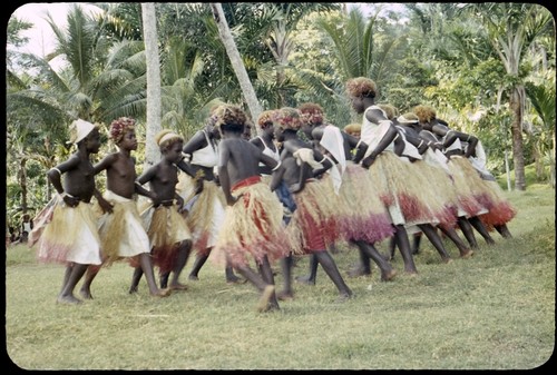 Dancers in grass skirts and matching headress; some with western dress or tops, and white handkerchiefs tied around head or arm