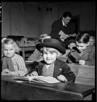 [First French Lesson: small child, Hans, in dark (Alsatian?) suit and round hat, in school room with older children]