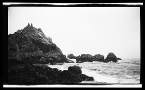 Large rocks along the ocean, showing two people at the peak of the tallest rock with one person slightly raising arm