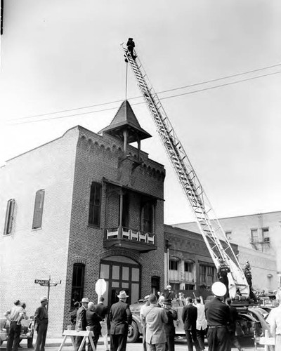 City Fireman John Reeves, Truck Company No. 20, is shown above on an aerial ladder affixing a "No. 1" to the helmet which is on top of the flag pole on Los Angeles's fire firehouse, located at Plaza and Los Angeles Streets