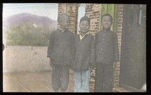 Three smiling boys standing near the entrance to a brick building, China, ca. 1918-1938