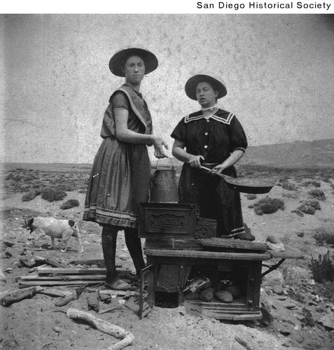 Two women in bathing suits working over a wood cooking stove