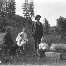 Exterior view of a family of five, some seated out in a field. Family posing in a garden with chickens, a man in a barber chair, and children playing