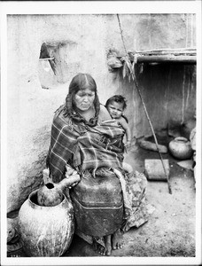 Hopi Indian mother with her baby on her back getting water from an olla outside an adobe dwelling, ca.1900