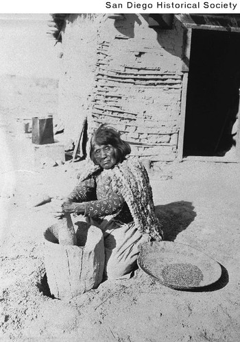 Unidentified Indian woman grinding grain while kneeling on the ground outside an adobe shack