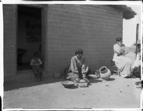 Family outside adobe structure. Woman with metate