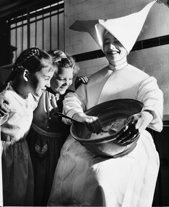 Two girls of the Los Angeles Orphanage watch Sister Elizabeth mix cake batter, 1951