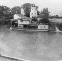 Sacramento River, 1906. by H. Douglas. Photo of a small steamship along river bank with a large tankhouse and other buildings visible