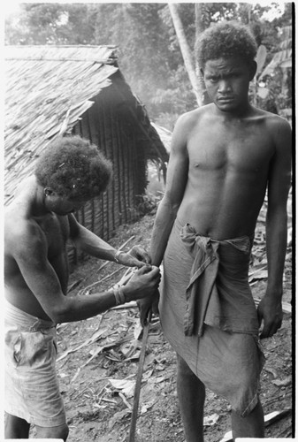 Man ties red cane obi on another man's wrist prior to a feast they are going to give
