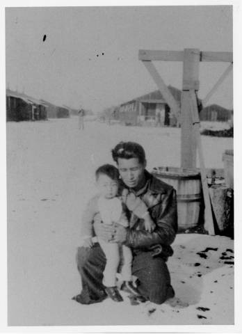 Man with small child on his knee outside [in snow?] with barracks of Tule Lake Relocation Center in background